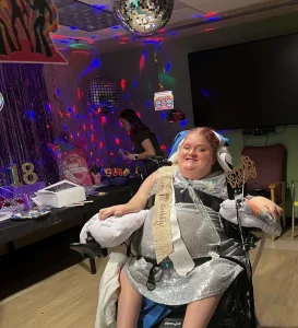 Young disabled girl dressed up for her birthday party, in a wheelchair, with party decorations in the background.